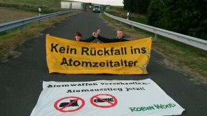 Zwei Aktivist*innen und zwei Banner ("Kein Rückfall ins Atomzeitalter" und " Atomwaffen verschrotten - Atomausstieg jetzt! Robin Wood") vor Tor 1 des Fliegerhorstes in Büchel